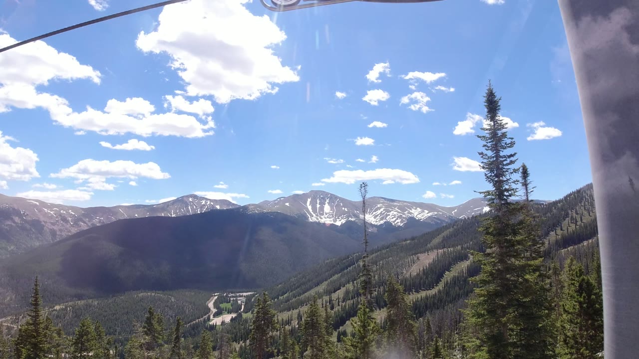 Wide Winter Park, Colorado mountain panorama with evergreen slopes, a tall foreground conifer at right, distant peaks with snow patches, and no visible people.
