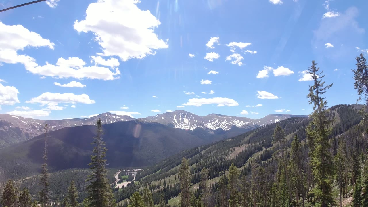 Wide mountain valley with pine trees and snow-streaked peaks under blue sky.