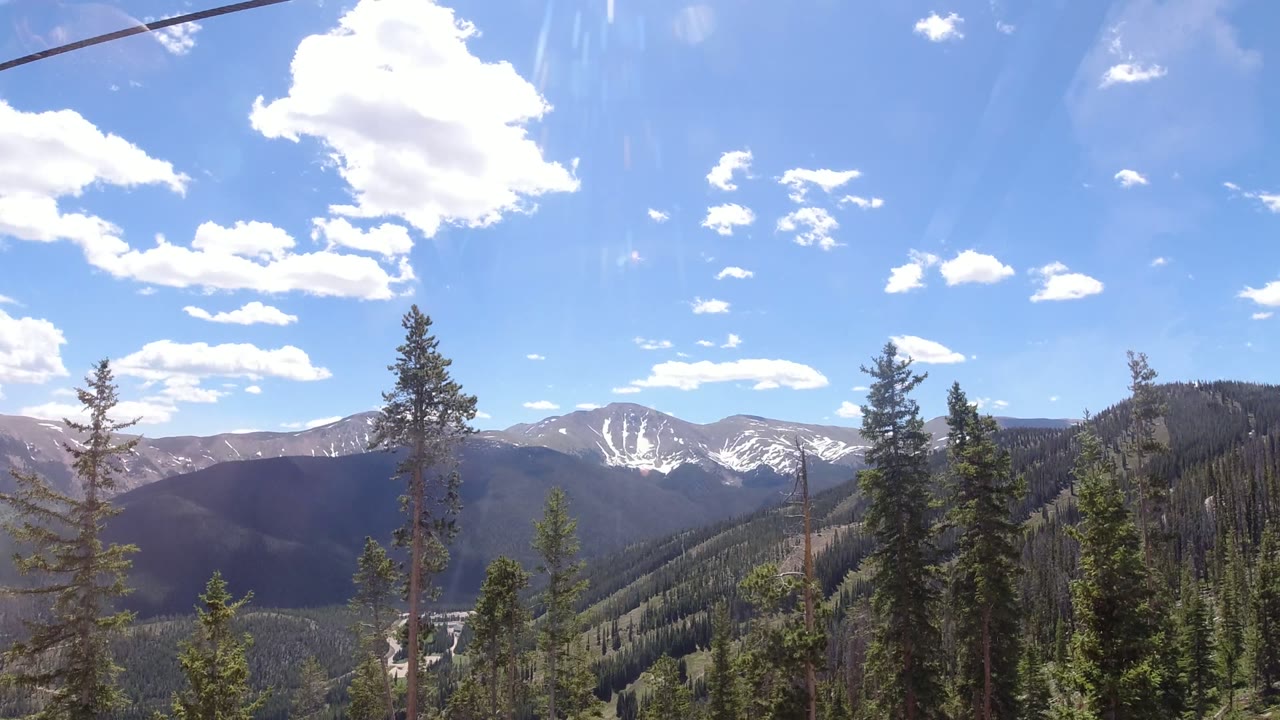 Wide mountain view with evergreen slopes, ski lift cable, blue sky, and distant snow patches.
