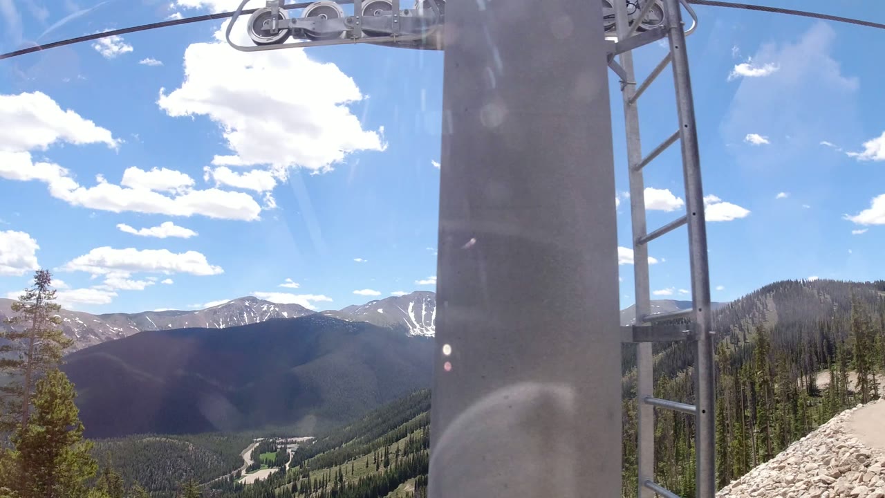 Winter Park, Colorado ski lift tower and cable hardware pass close to frame, with layered mountains, evergreen forest, snow patches, and no visible people beyond it.