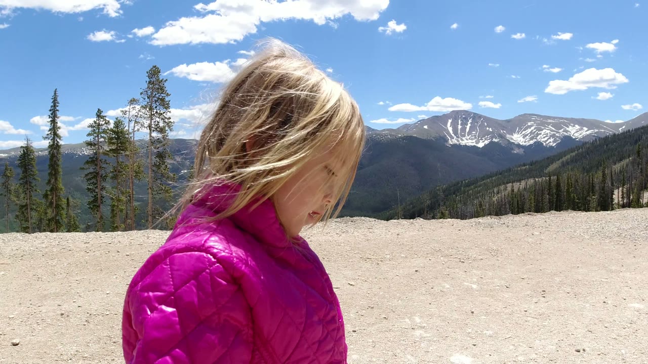 The girl appears closer to center frame in Winter Park, Colorado, her hair blown across her face as pale rock, dark forested ridges, snowy peaks, and bright clouds remain visible behind her.