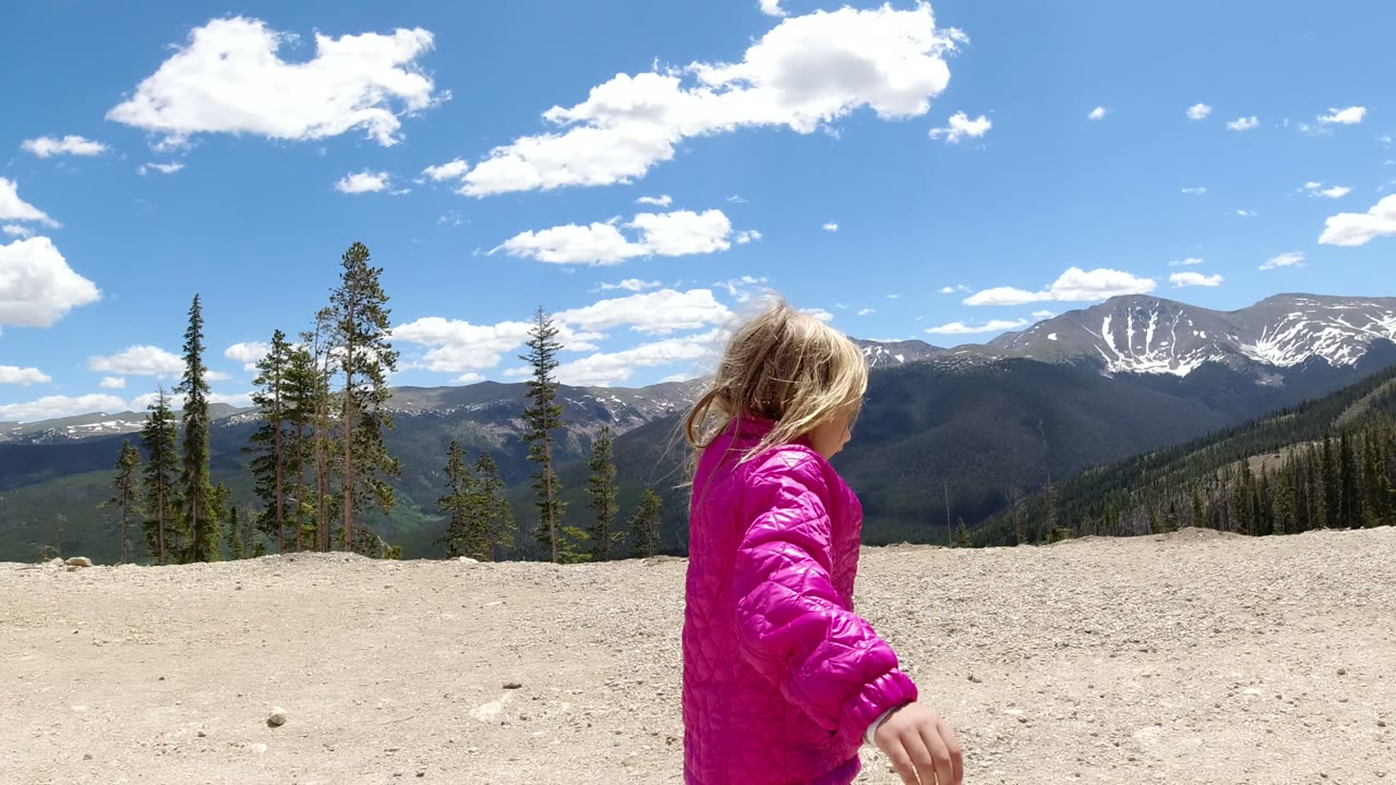 Young girl in a pink jacket walking along a mountain ridge with snowy peaks behind her.