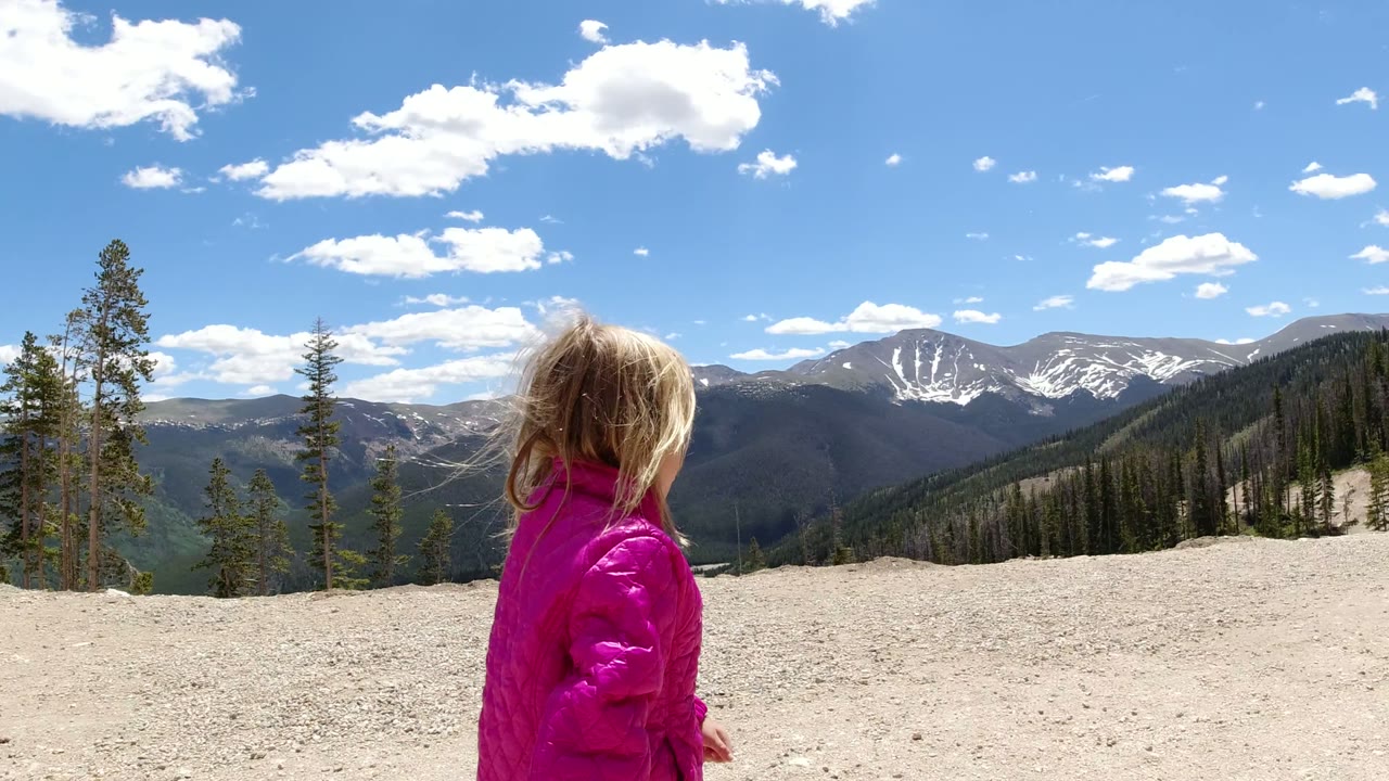 One second in, the girl is closer to center frame with wind-tossed hair, looking across the Winter Park, Colorado mountain ridge toward dark slopes and snow patches.