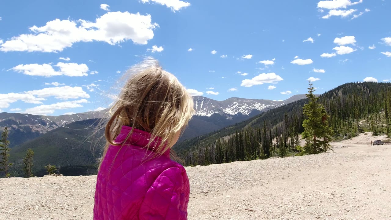 The child faces away from the camera on a sunlit dirt path in Winter Park, Colorado, with dark green forested slopes and distant snowy peaks under scattered clouds.