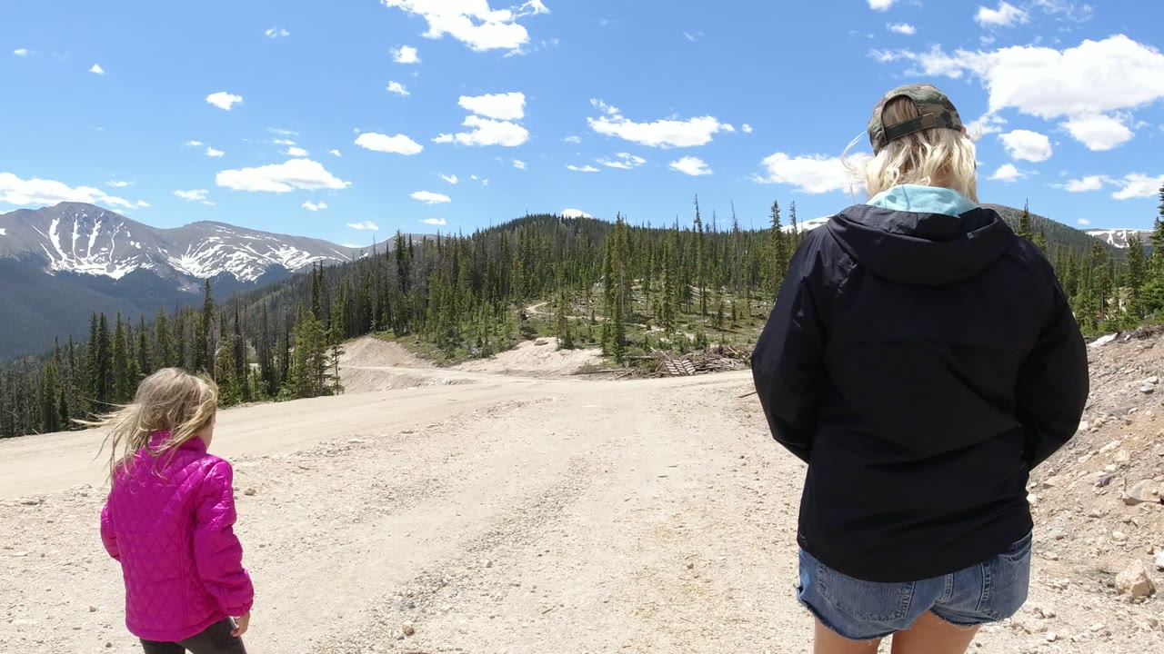 In Winter Park, Colorado, the child walks near the left side of the gravel road while the adult is partly cropped on the right, with evergreens and distant mountain ridges under bright sun.