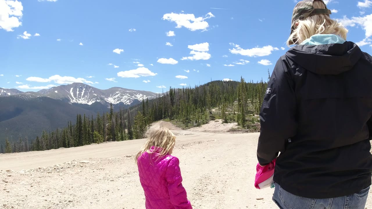 In Winter Park, Colorado, a blonde adult in a dark jacket and cap walks behind a small child in a pink coat toward a distant snow-streaked mountain range.