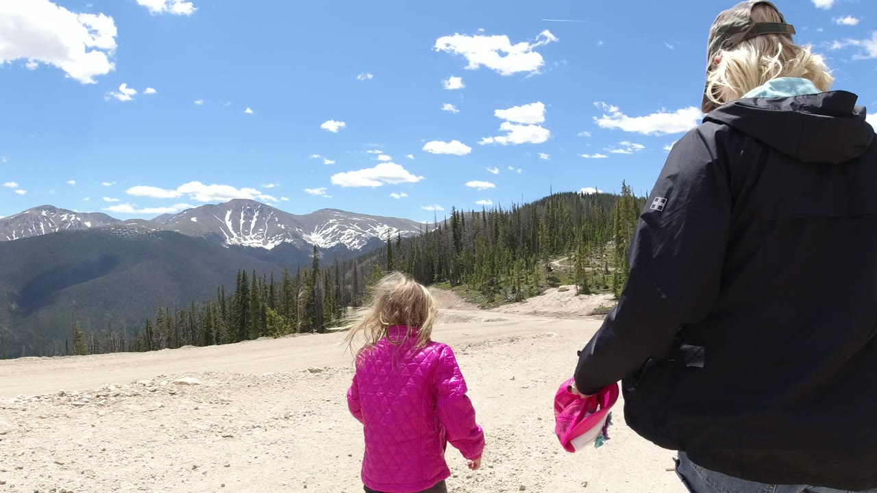 The child in a bright pink coat moves farther into the open foreground while the adult remains on the right side, with Winter Park mountain ridges and pine trees beyond.