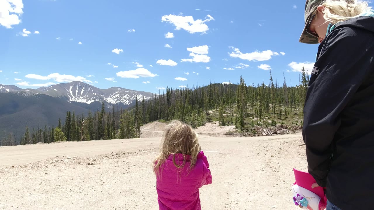 A young child in a bright pink coat is centered low in the frame while a woman is partly visible at the edge, with forest and snowy peaks beyond in Winter Park, Colorado.