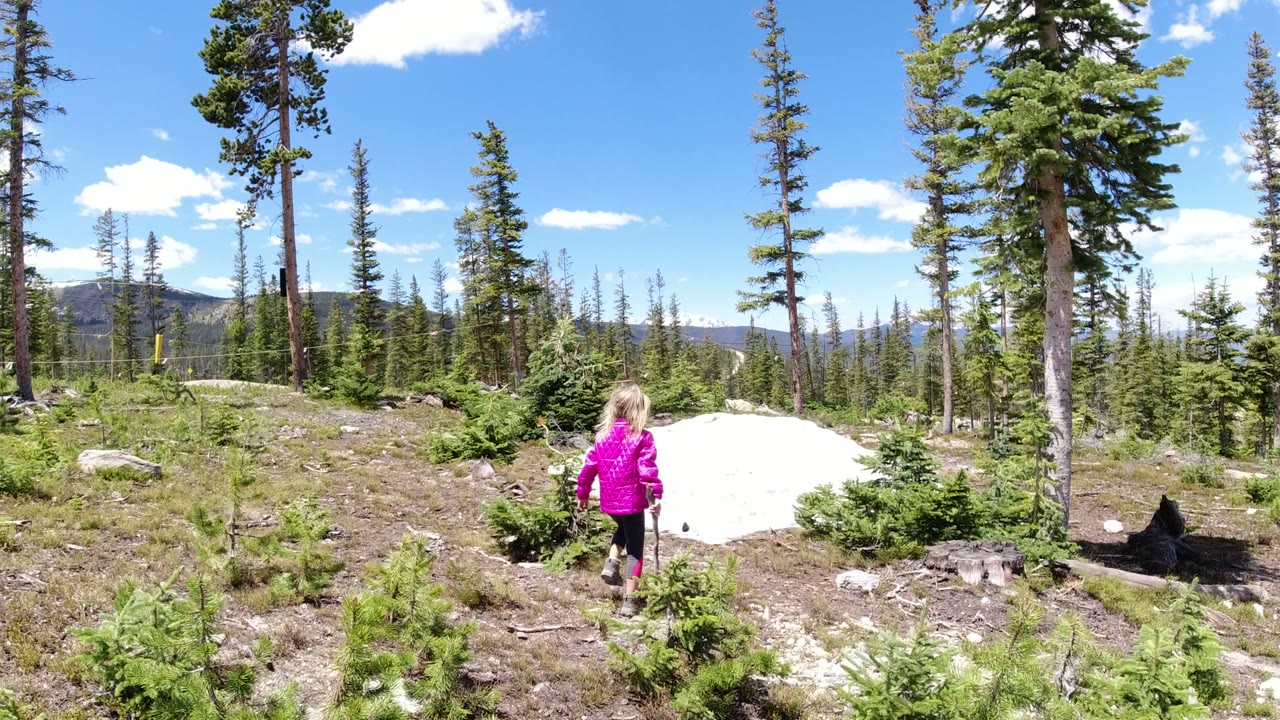 The girl stands closer to the center of the Winter Park, Colorado clearing, holding a stick near low evergreens and a bright white snow patch.