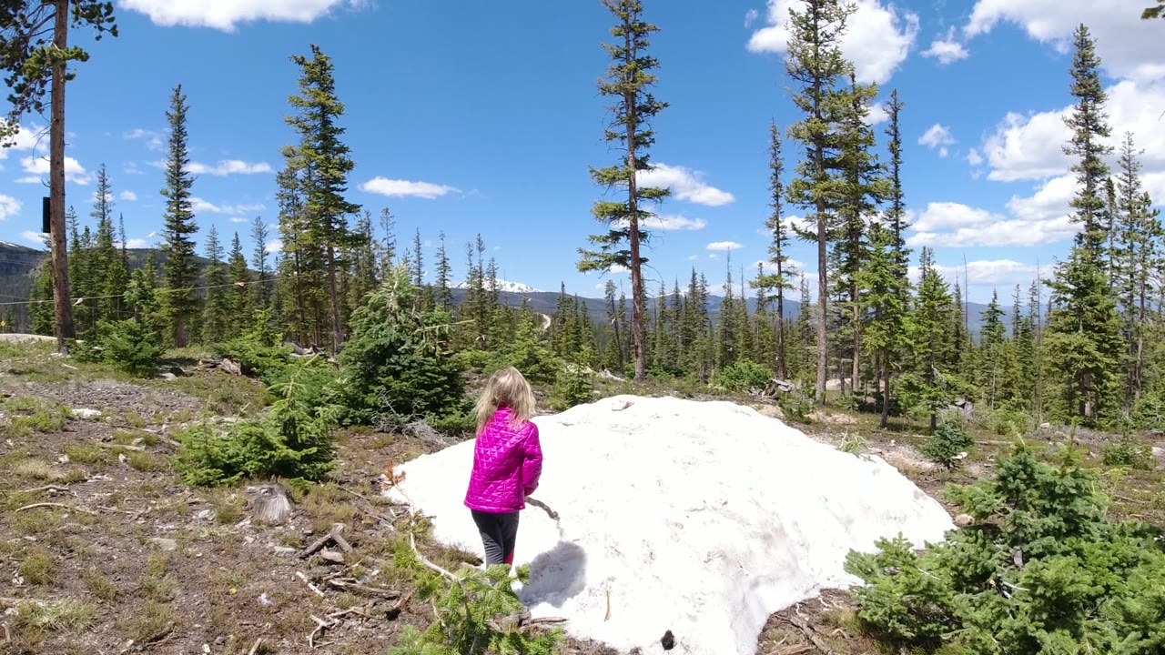 One second in, the girl stands closer to the snow patch in Winter Park, Colorado, with small evergreen shrubs, bare ground, and tall trees filling the frame.