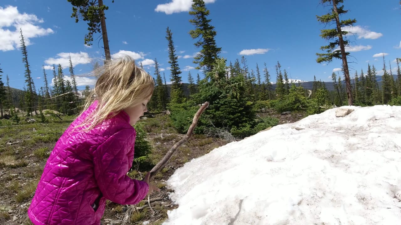 At the start of the Winter Park, Colorado clip, a blonde child in a bright pink jacket holds a stick near a white snow or rock-covered mound with evergreen trees and blue sky behind her.