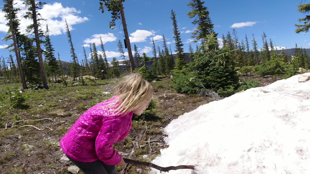 One second in, the child leans forward with the stick angled toward the white mound in the Winter Park, Colorado wooded landscape.