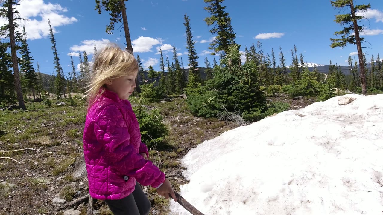 A child in a bright pink jacket stands at the left edge of a Winter Park, Colorado forest clearing, holding a stick beside a white snowbank under blue sky.