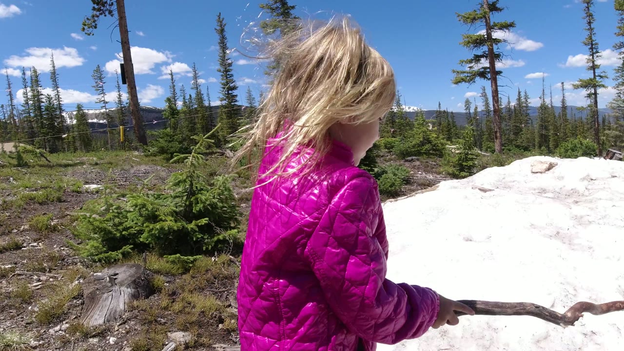 The child in the pink jacket is closer to the center-left of the frame in Winter Park, Colorado, holding the stick beside the snow patch with evergreens and distant hills behind.