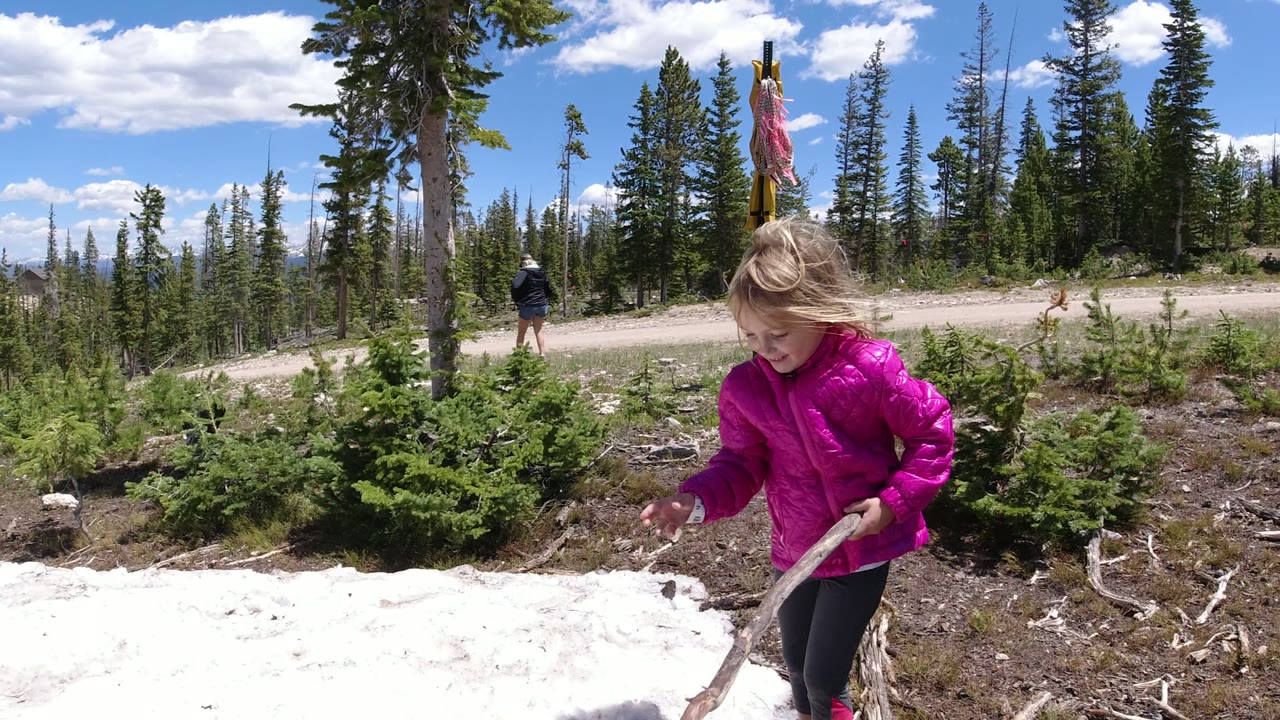A young girl in a bright pink jacket holds a stick near a snow patch in Winter Park, Colorado, with pine trees and a dirt road behind her.