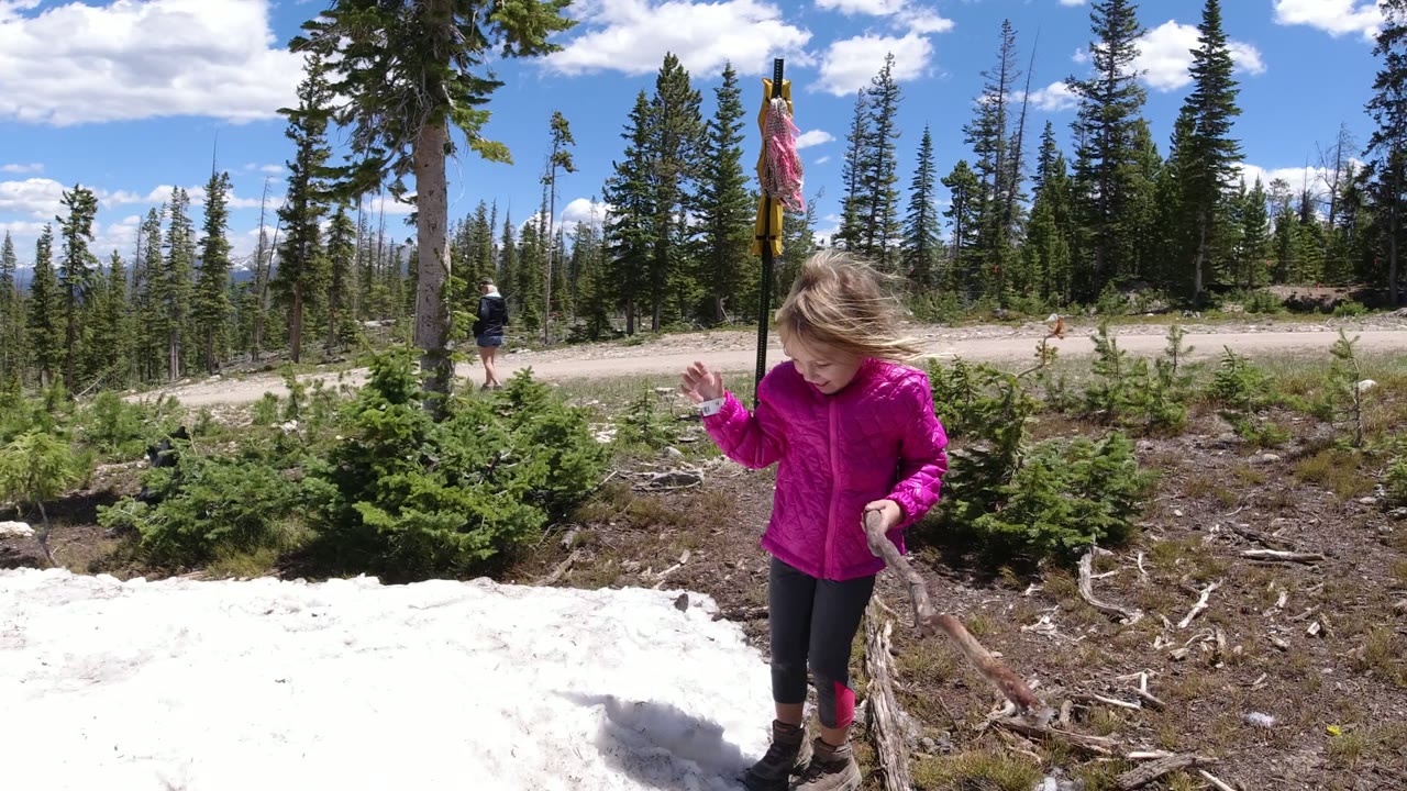 The child stands near the right side of the snow patch in Winter Park, Colorado, with one hand raised, a stick in the other hand, and tall pines in the background.
