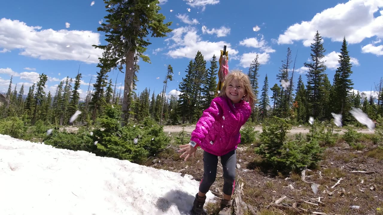 The girl smiles toward the camera as pieces of ice scatter in front of a snowy slope and pine trees in Winter Park, Colorado.
