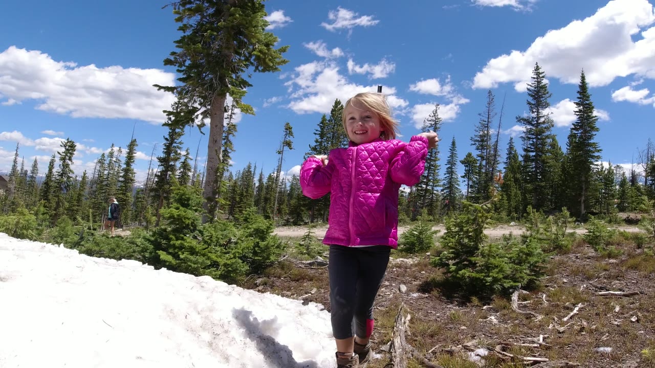 Blonde child in a bright pink jacket smiles beside a snowbank in Winter Park, Colorado, with pine trees and a distant adult behind her.