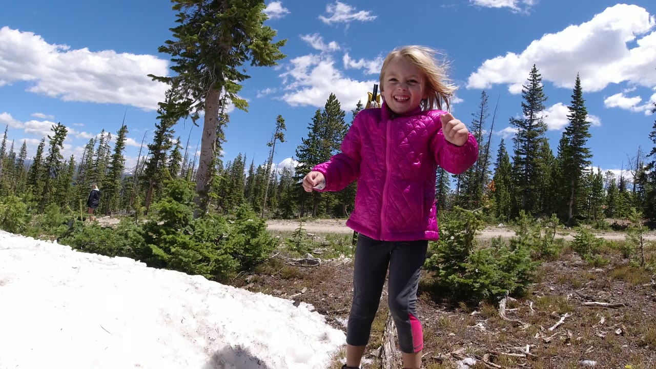 The child stands closer to camera in Winter Park, Colorado, smiling in a pink jacket with a white snowbank, scrubby ground, and tall pines behind her.