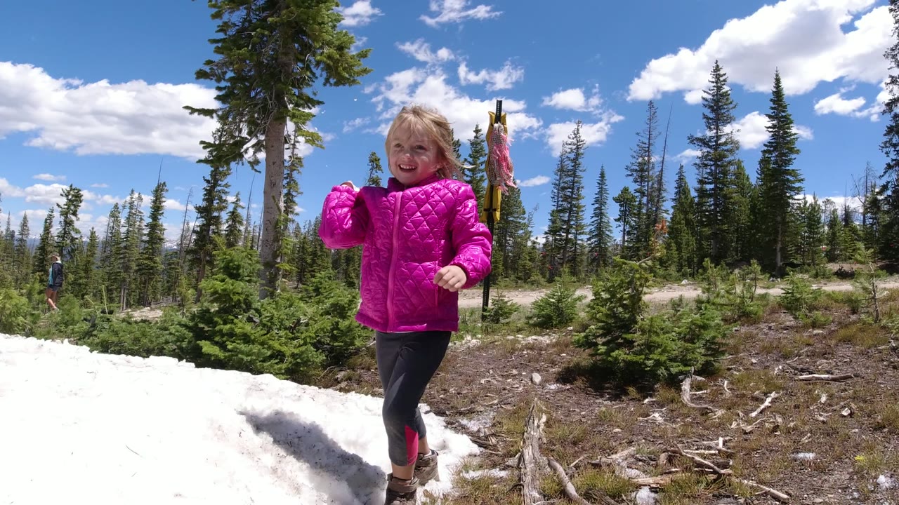 A young girl in a bright pink jacket stands near a snow patch in a Winter Park, Colorado forest, with conifers, blue sky, and a distant adult figure in the background.