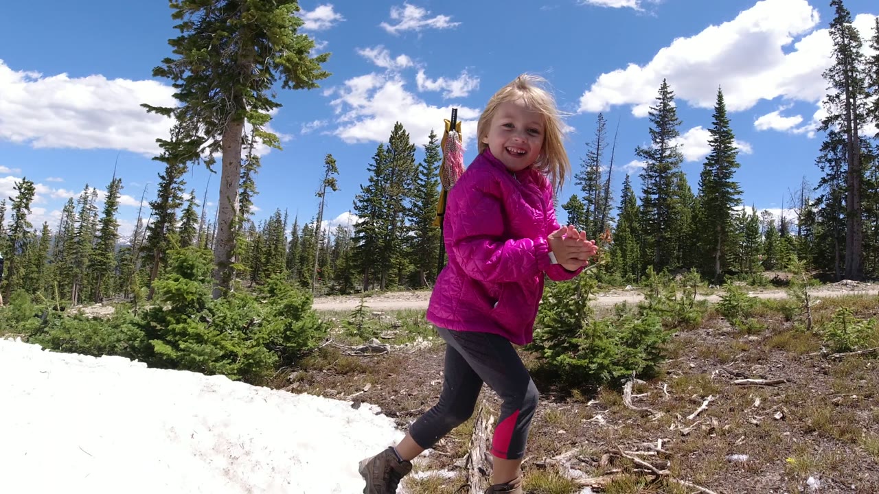 The young girl smiles while stepping along the edge of a snow patch in Winter Park, Colorado, with small evergreens and tall conifers behind her.