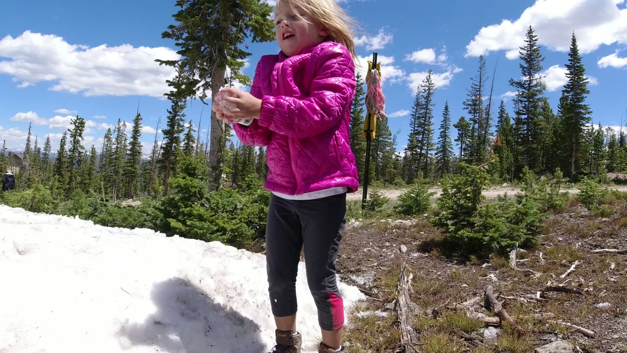 The child stands close to the camera beside a snow patch in Winter Park, Colorado, holding snow with trees and open sky in the background.