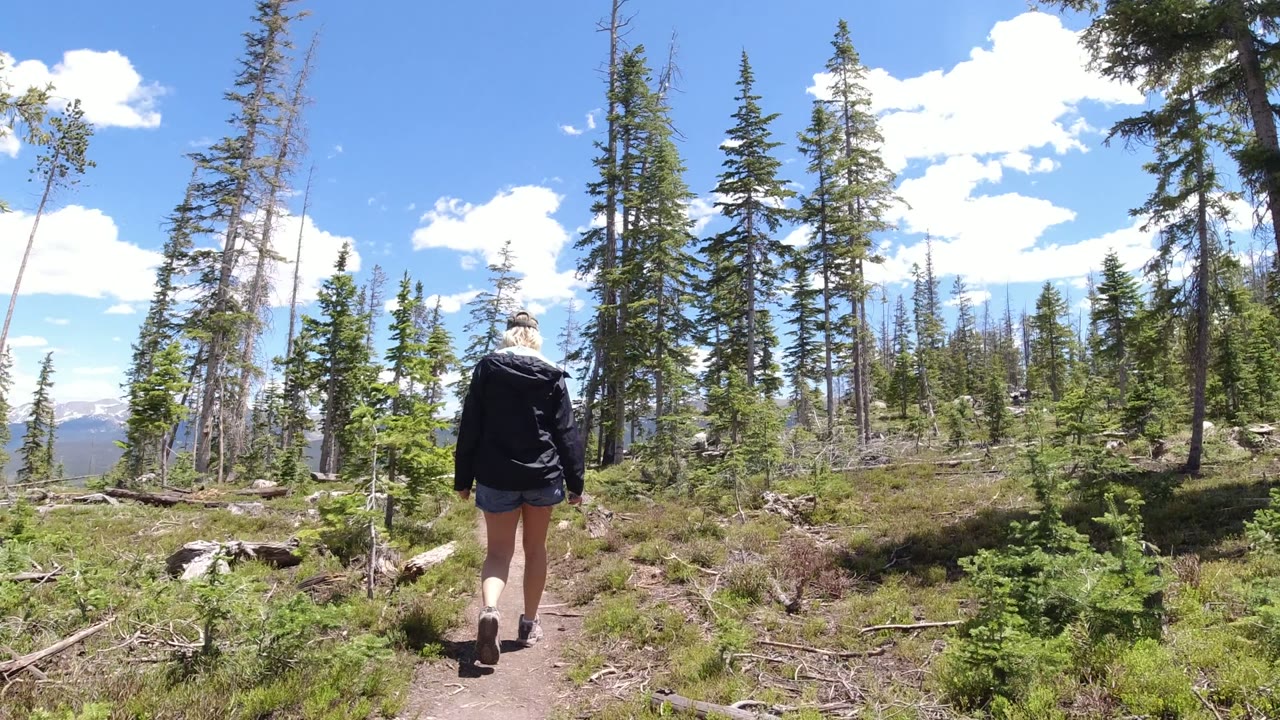 At 0:00, a woman walks away along a narrow dirt trail in Winter Park, Colorado, with scattered evergreens, fallen branches, blue sky, and distant mountains visible through the trees.