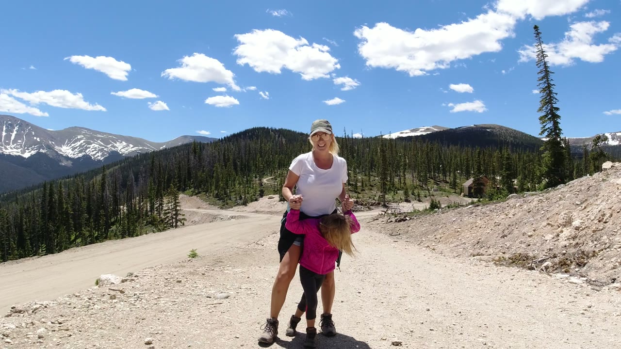 The woman and young girl stand closer in the foreground on the Winter Park dirt road, with the child leaning forward and the mountains still visible over the tree line.