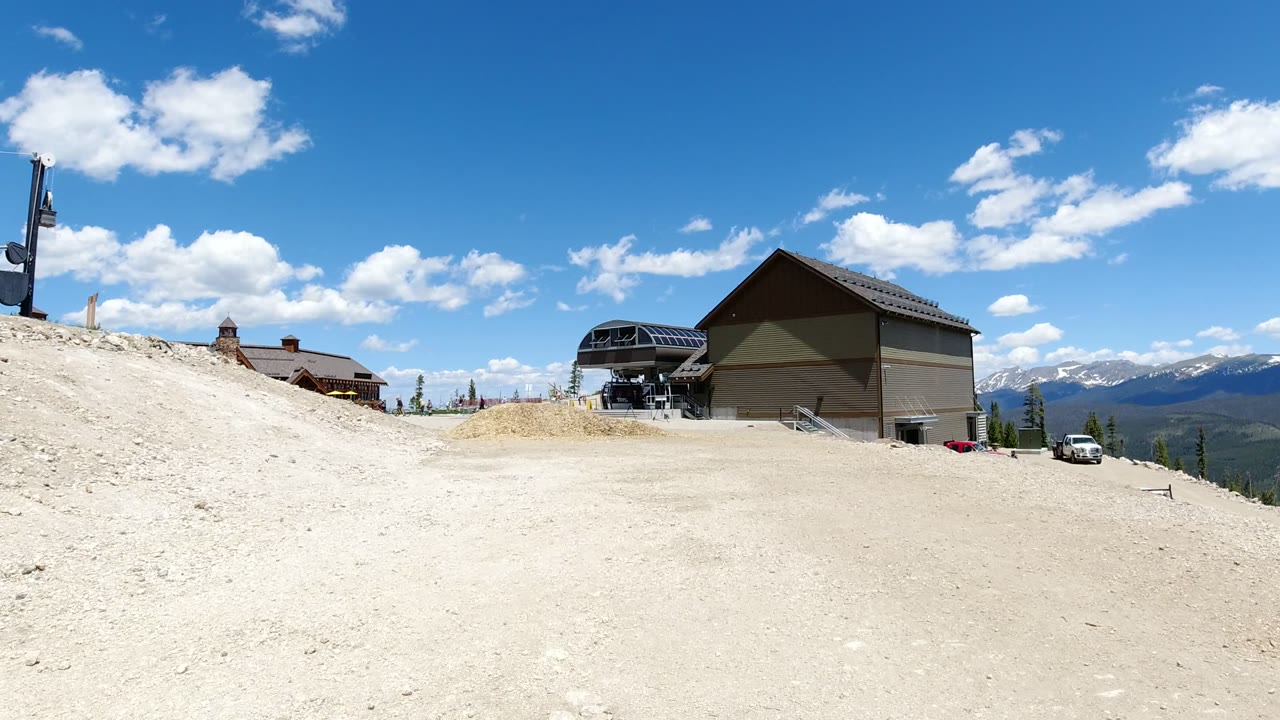 Wide view of gravel ground, mountain buildings, lift terminal, clouds, and distant peaks.