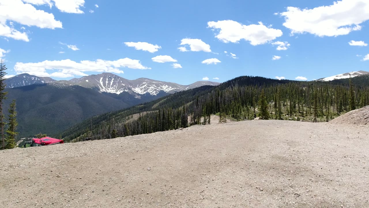 Wide mountain overlook with gravel foreground, pine trees, distant snow-streaked peaks, and blue sky.