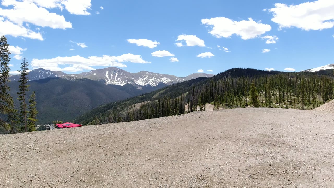 Winter Park, Colorado mountain panorama with gravel across the lower frame, pines near the ridge, layered dark hills, snow-streaked peaks, and no visible people.