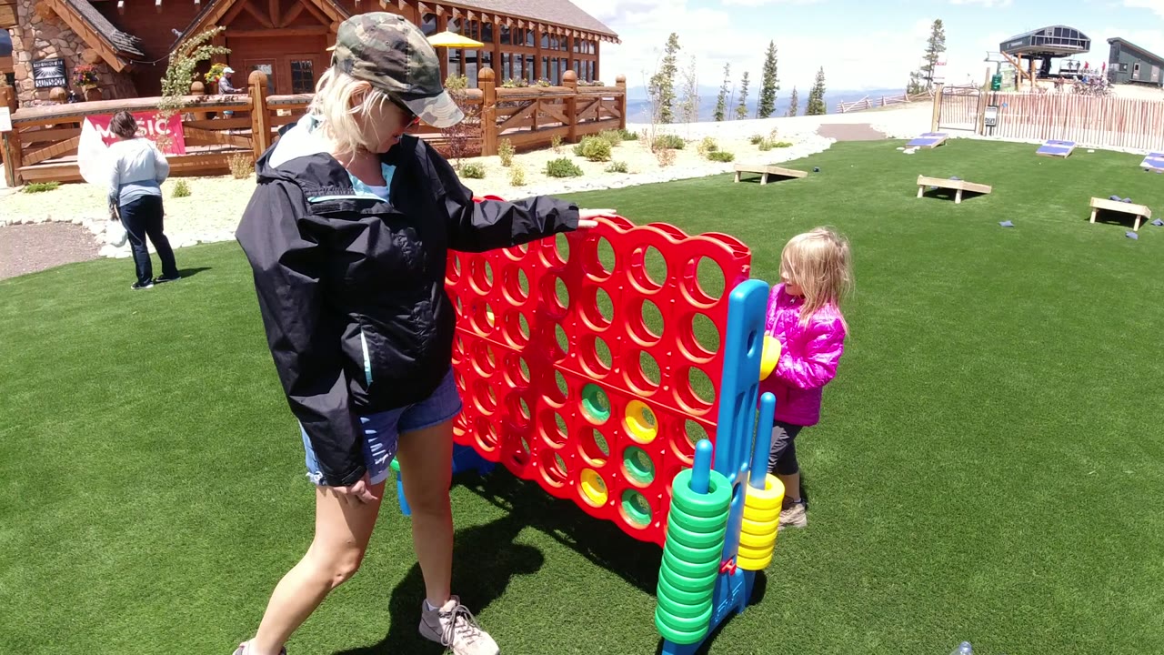 At Winter Park, Colorado, a woman in a cap stands next to a large red connect-four style game while a young girl in a pink jacket faces the board on green turf.