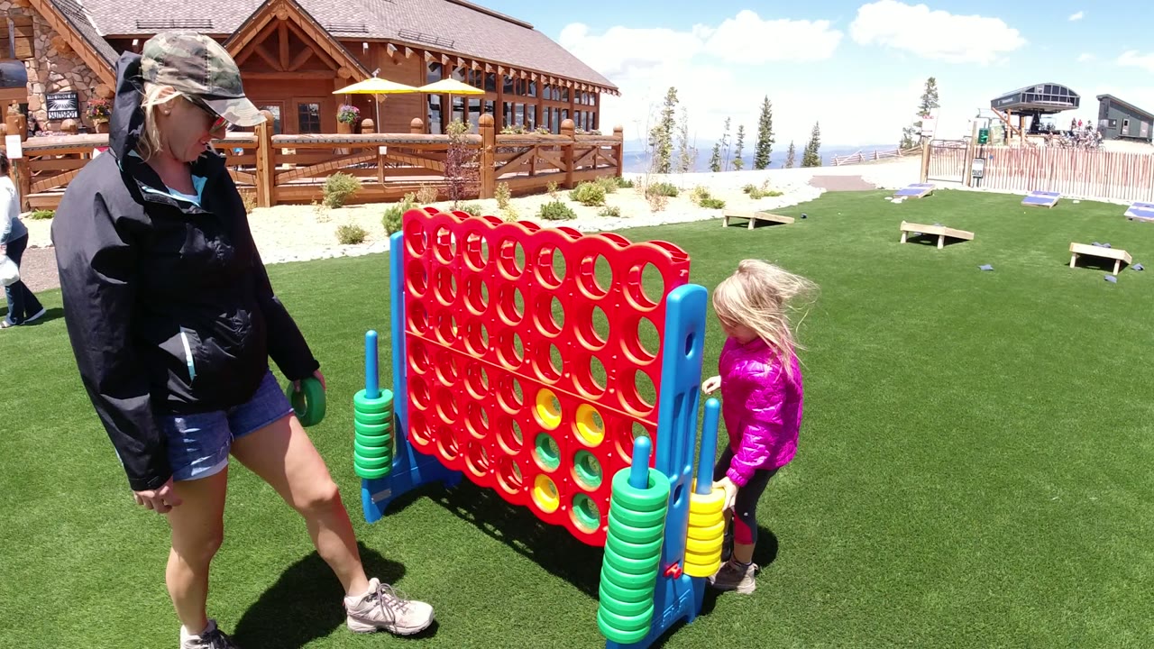 In Winter Park, Colorado, a woman in a cap stands near an oversized red Connect Four board while a young girl in a pink jacket waits on the opposite side.