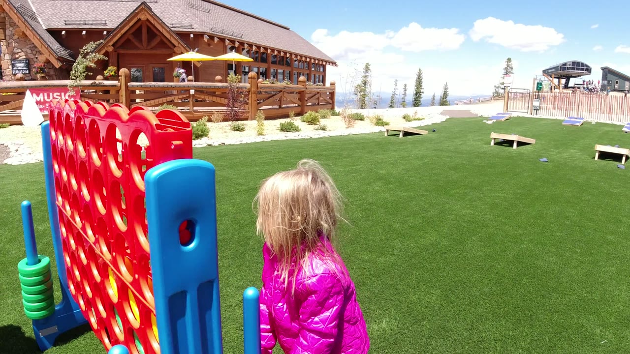 The child in a pink jacket faces the oversized red game board on the lawn in Winter Park, Colorado, with cornhole boards, a stroller, fencing, trees, and a lodge behind her.