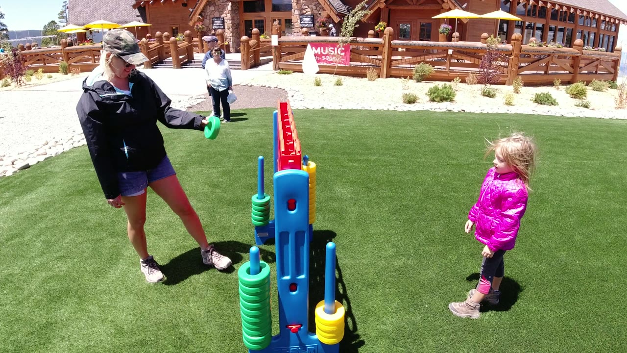 The adult lifts a green disc toward the tall game board as the young girl stands to the right on the bright turf in Winter Park, Colorado.