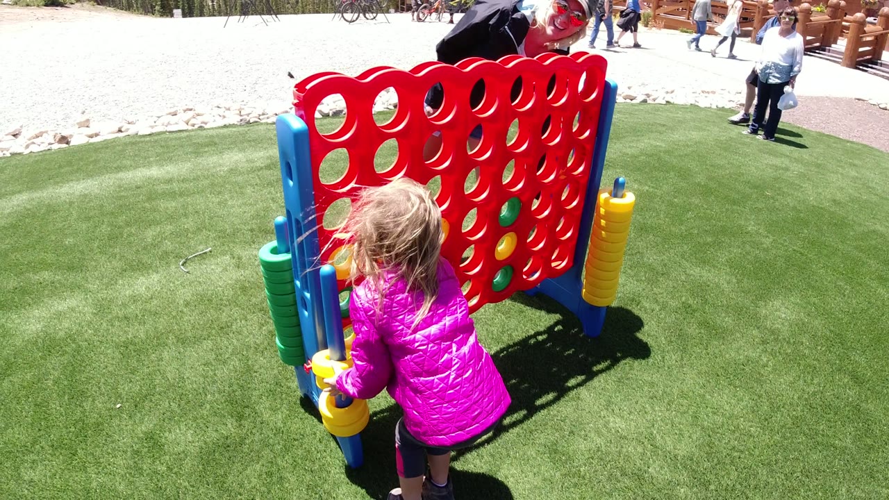 The girl moves to the blue side support of the oversized Connect Four game in Winter Park, Colorado, with stacked yellow and green pieces visible beside the board.
