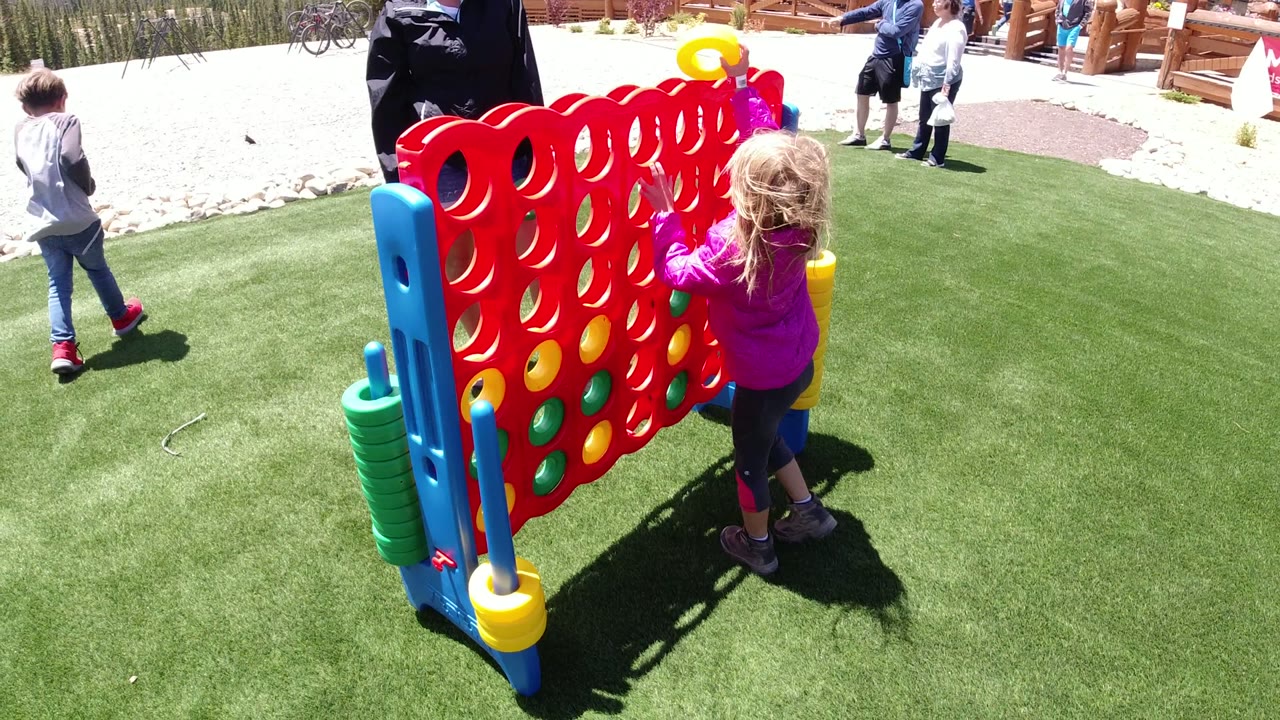 Young girl in a purple jacket plays beside a large red game board on green turf.