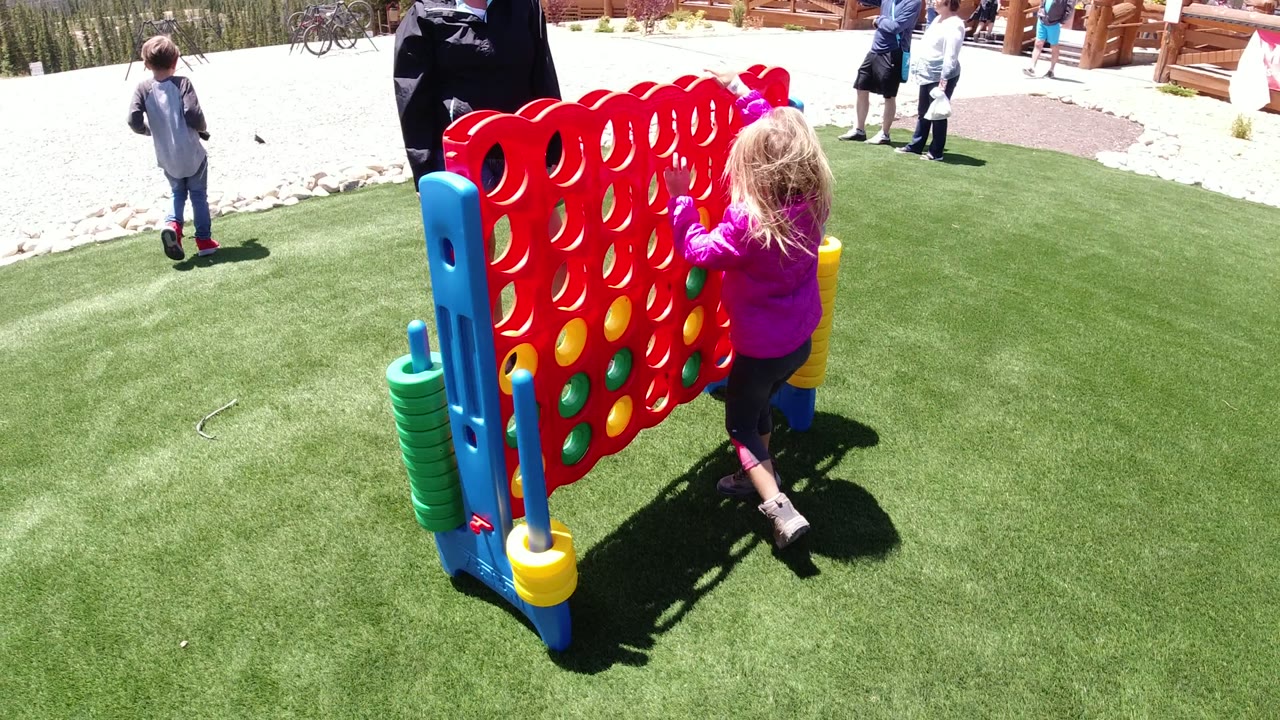 At 0:01 in Winter Park, Colorado, the girl remains beside the large red game board, with colored discs stacked on the side posts and visitors visible near the fence behind her.