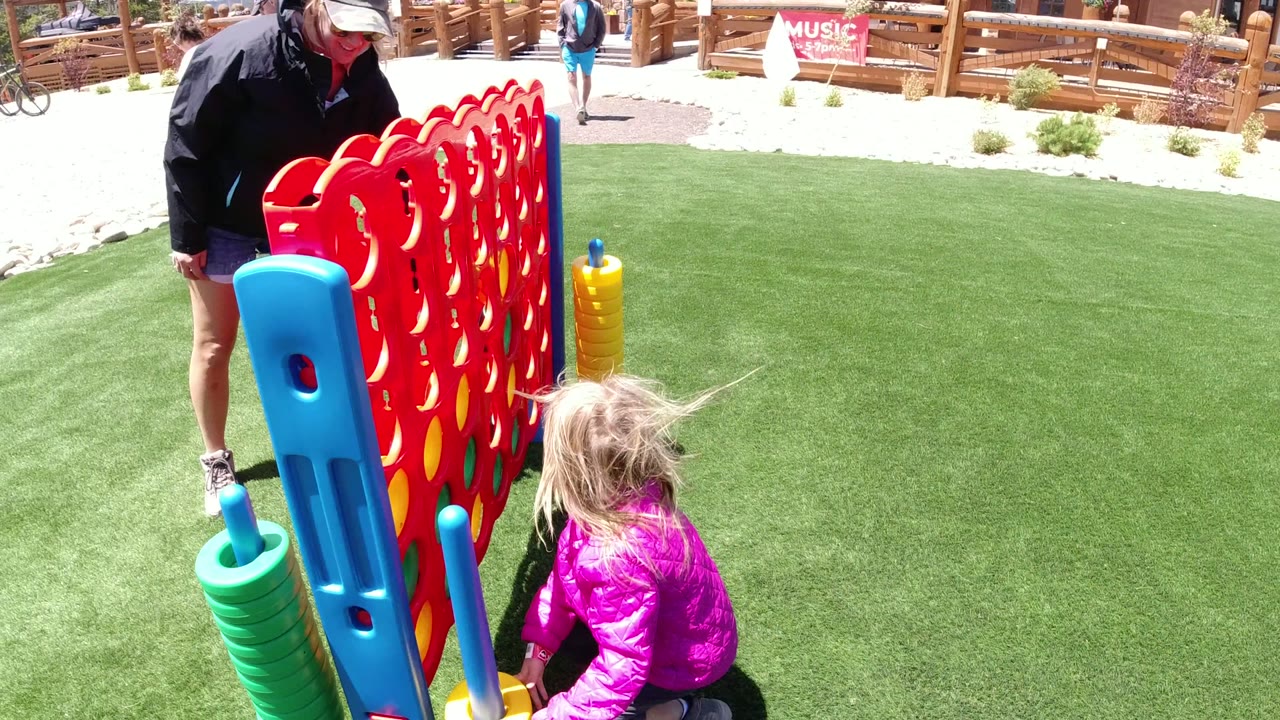 The child crouches at the side of the oversized red game board in Winter Park, Colorado, with stacked green pieces and a partial adult figure near the frame edge.