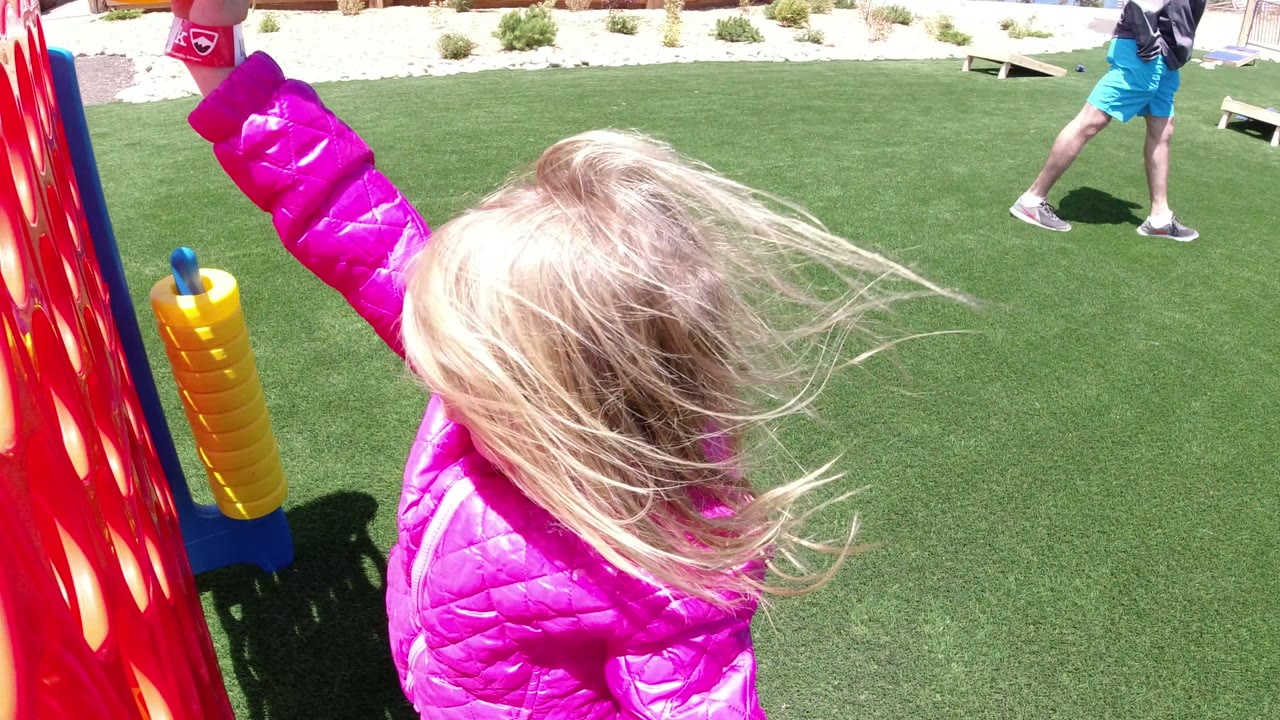 A blond child in a bright pink coat stands close to a red Connect Four style game on green turf in Winter Park, Colorado, with another person partly visible in the background.