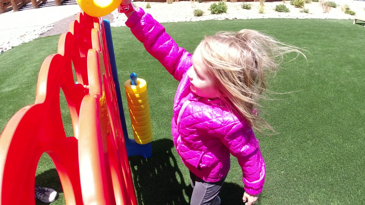 The child turns near the red game board in Winter Park, Colorado, with blond hair blown across her face and green turf filling most of the background.
