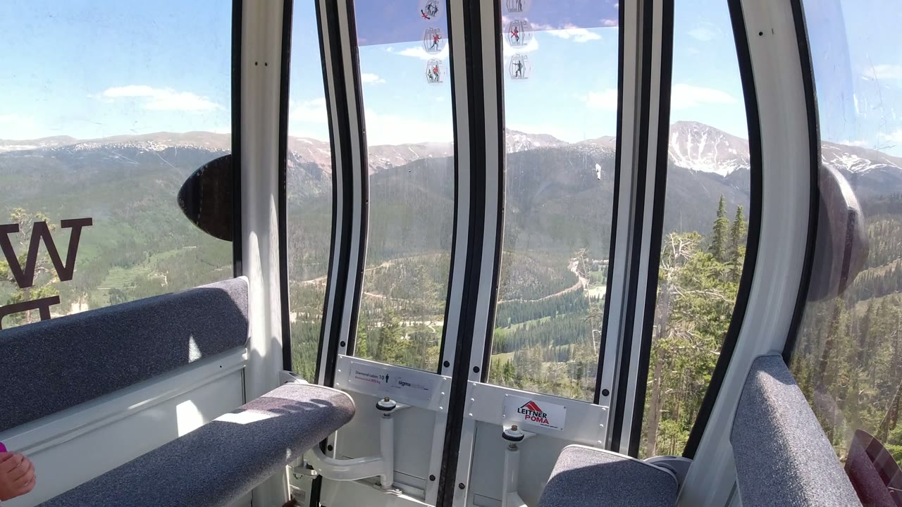 Second frame inside the Winter Park ride car shows gray seating and vertical window frames against a broad view of green mountain slopes and pale snowy peaks beyond.