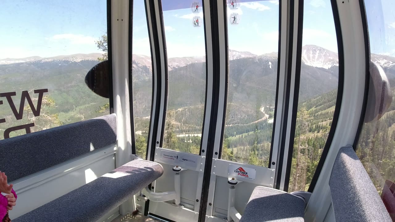 Lift cabin window view over forested mountains and snow-capped peaks.