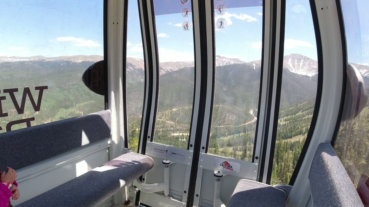 Opening frame inside a cable car in Winter Park, Colorado, with curved windows, gray bench seating, and green mountain slopes beyond the glass.