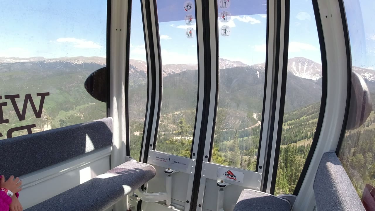 A second view from the Winter Park, Colorado cable car shows tall black window frames, a gray seat back, evergreen slopes, and pale ridges in bright daylight.
