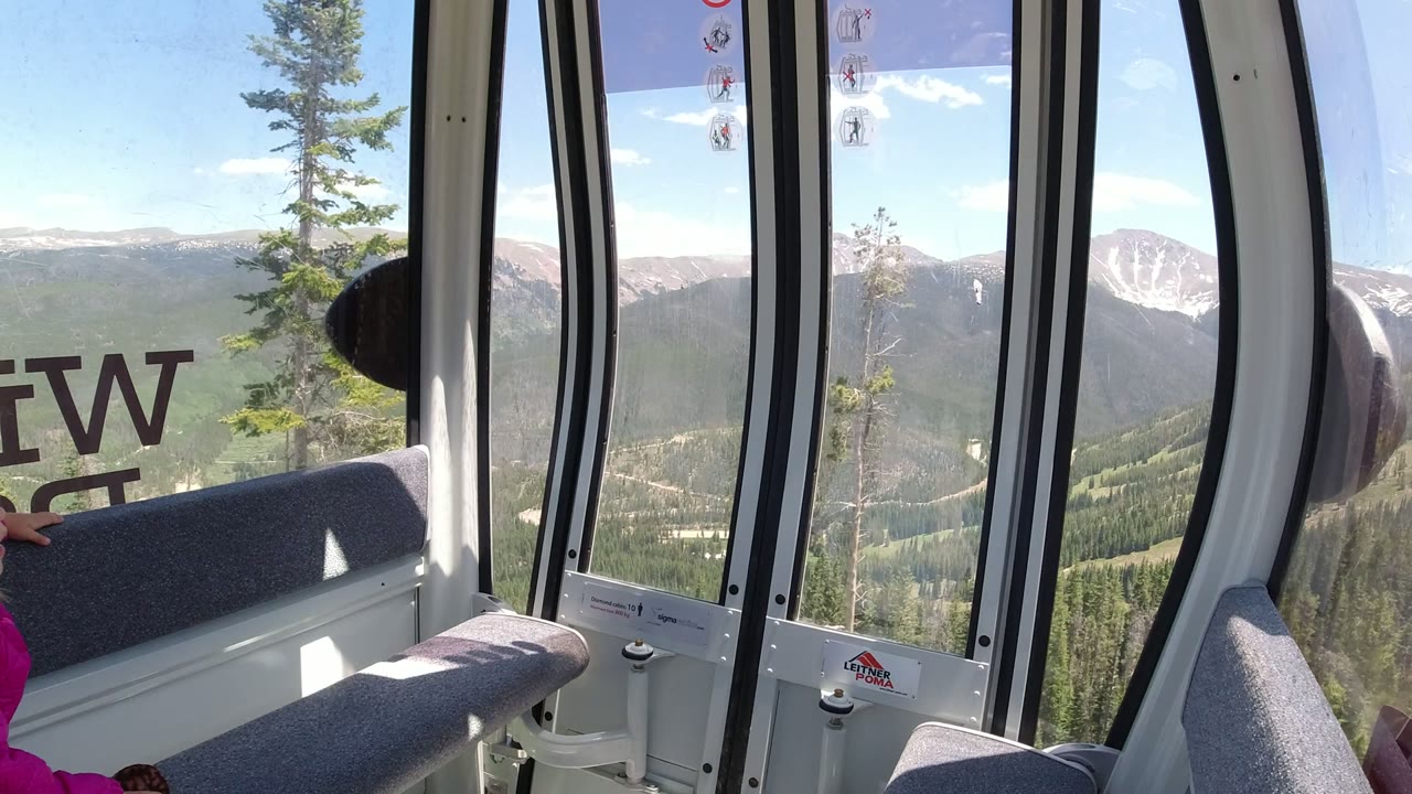 Inside a gondola cabin in Winter Park, Colorado, gray bench seats and tall windows frame pine-covered slopes and distant mountain ridges, with a partial person in pink at the left edge.
