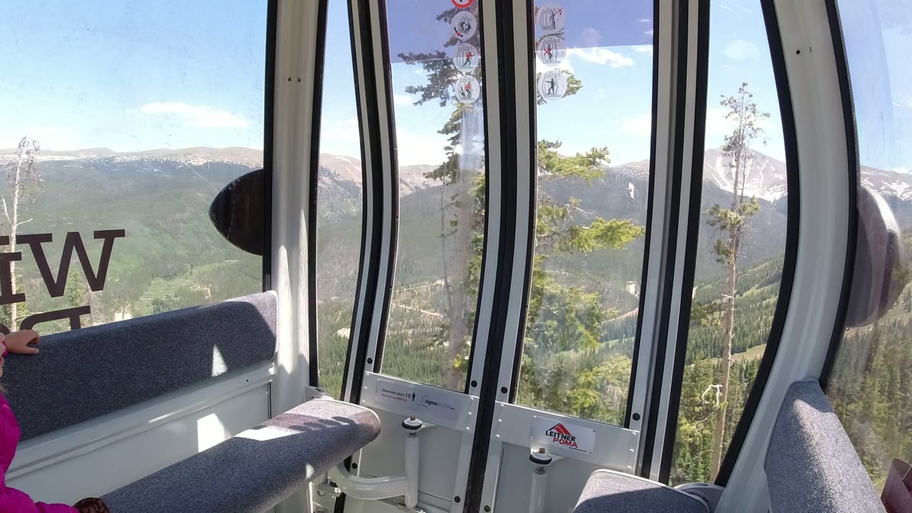 A Winter Park, Colorado gondola window view shows the bench, door frame, partial pink sleeve, forested valley, and pale mountain ridge under a clear blue sky.