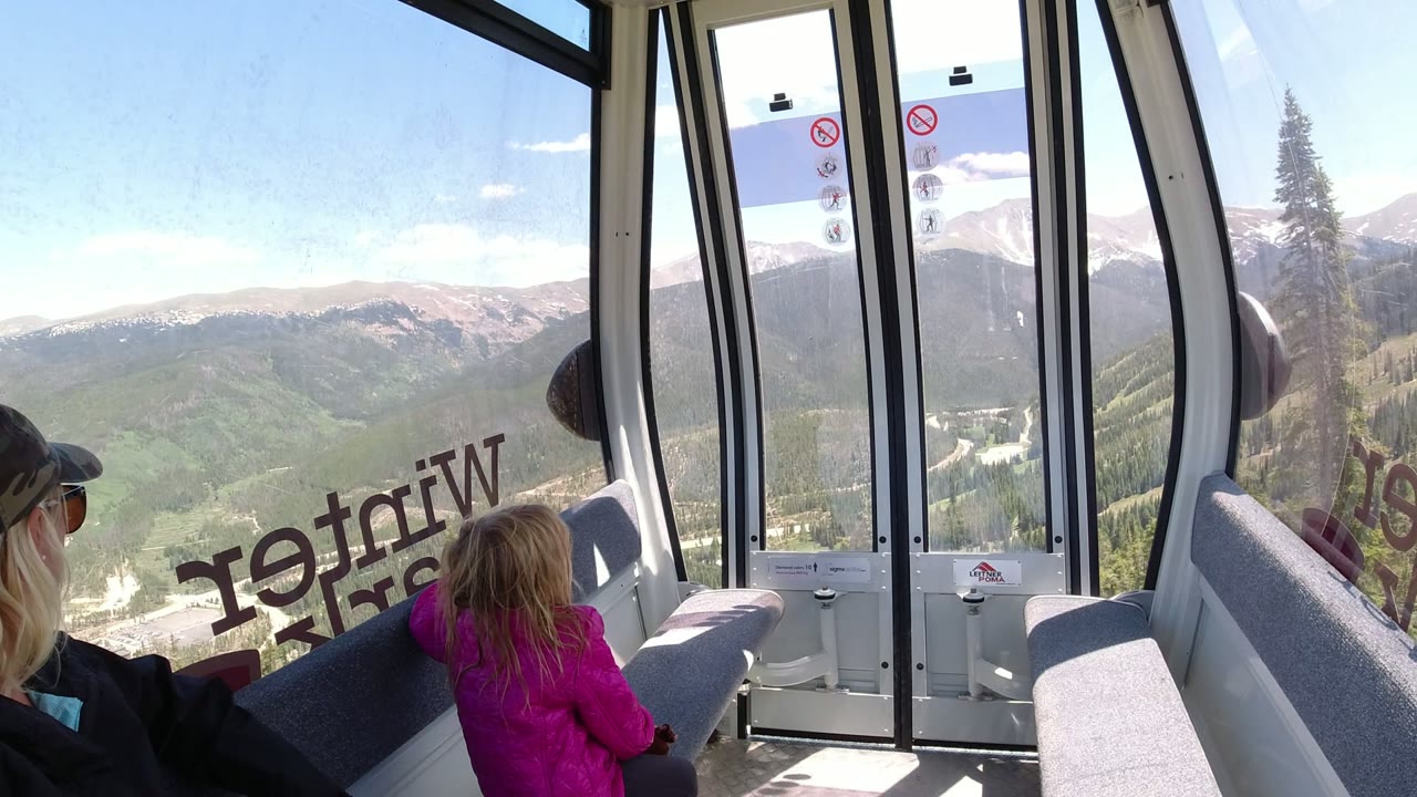 At 0:01, the seated woman and child remain low in the frame while the glass doors and vertical window bars frame the Winter Park mountain view outside.