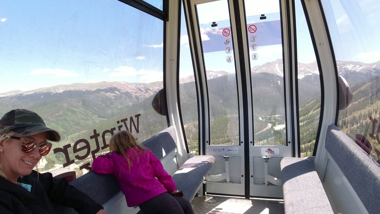 Woman in sunglasses and child inside a gondola with mountain views through the windows.