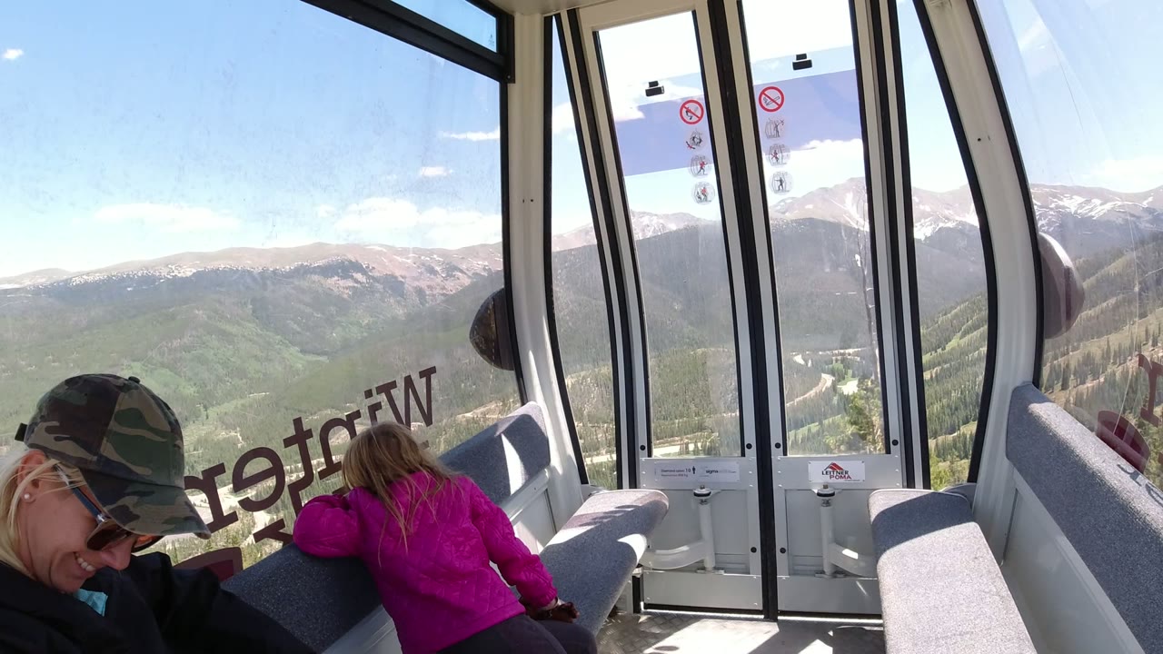 One second in, the child in a bright pink jacket remains near the gondola window as the seated adult is cropped at the left edge and Winter Park mountain ridges fill the background.